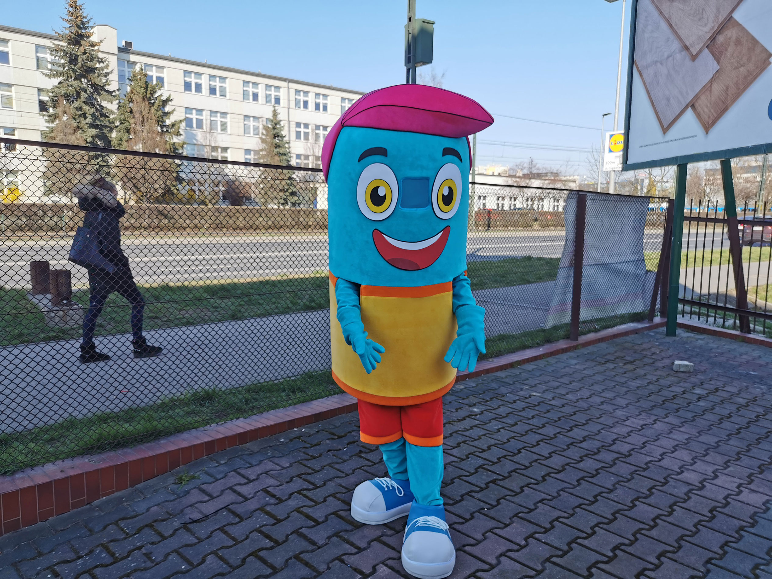 Na fotografii widnieje maskotka strój w oryginalnej postaci chłopca pozująca do zdjęcia przed siedzibą firmy Elabika